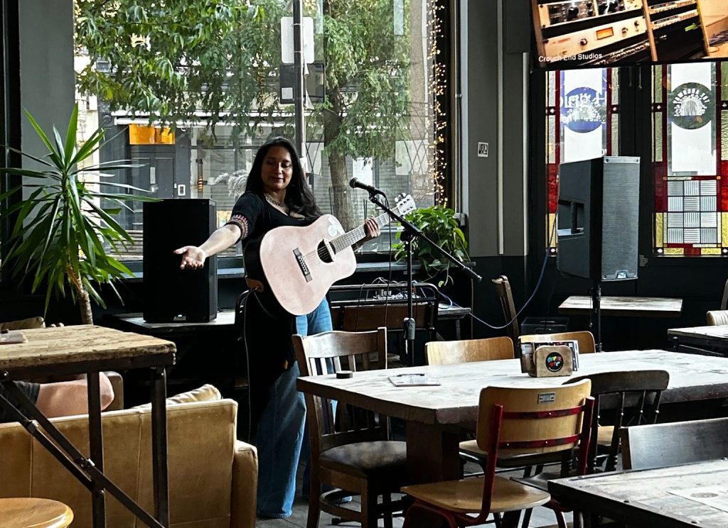 A musician performing with an acoustic guitar in a cafe setting, surrounded by tables and decorated with plants.