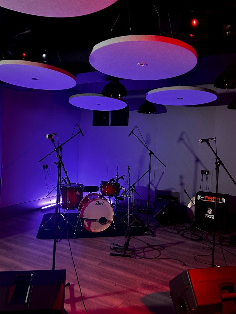 An empty stage setup featuring a drum kit and microphones in a dimly lit room with colorful lighting.